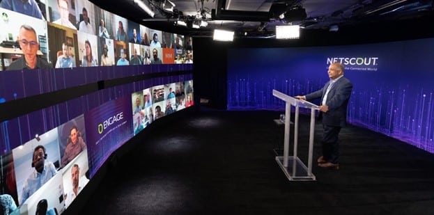Person at podium in a dim room with curved screen showing call participants; purple-blue theme and NETSCOUT logo on backdrop.