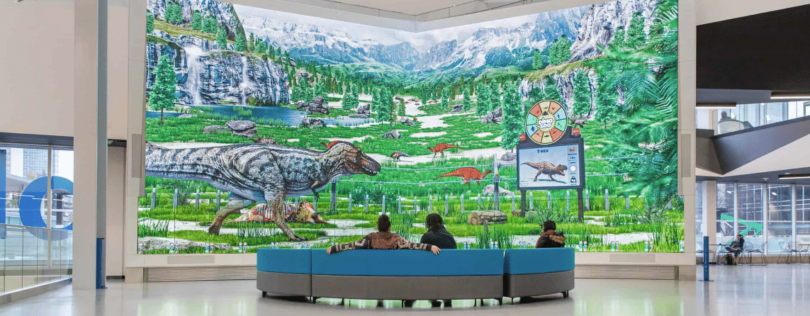 A large screen shows dinosaurs in a green landscape; three people on a circular couch watch, with mountains and waterfall behind.