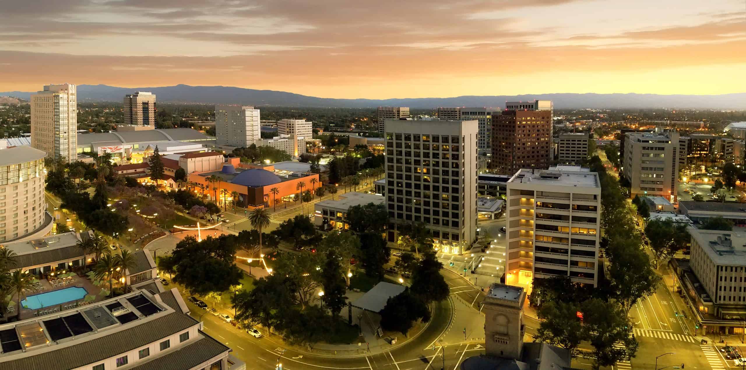 Aerial view: cityscape at sunset with lit buildings, streets below; distant hills and vibrant orange-yellow sky backdrop.