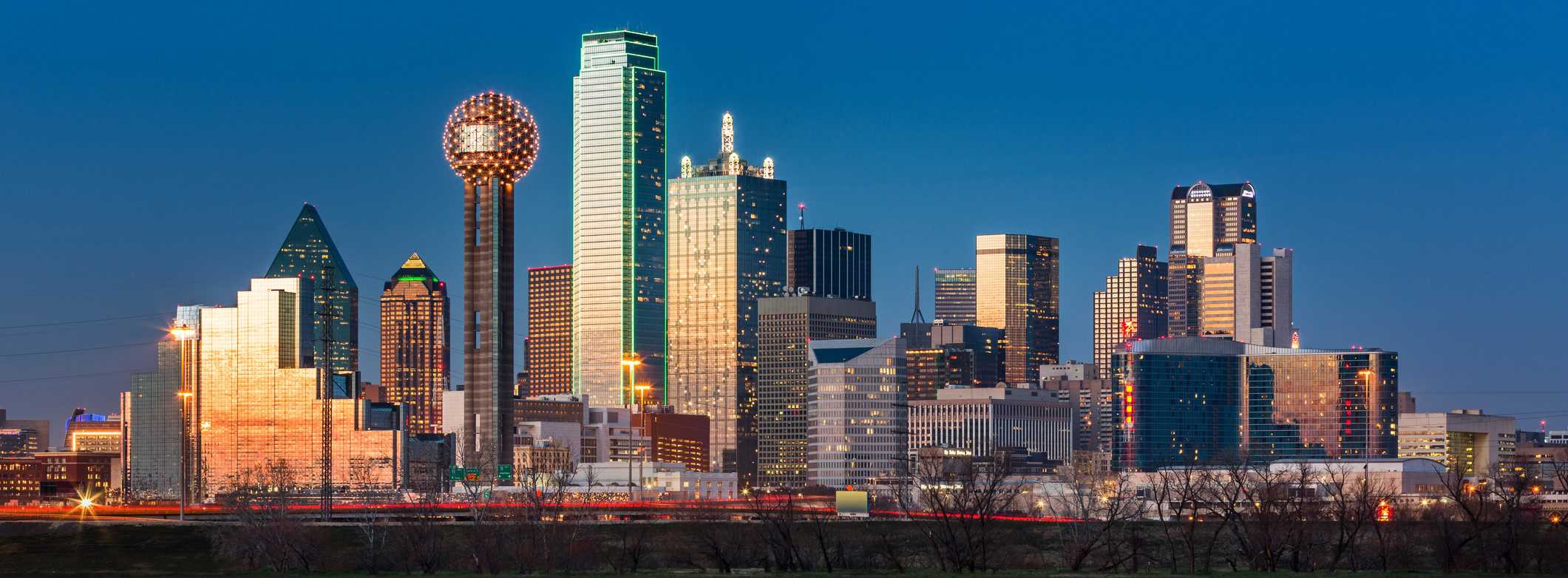 Dallas skyline at dusk: illuminated skyscrapers, Reunion Tower, clear sky, bare trees, and blurred taillights.