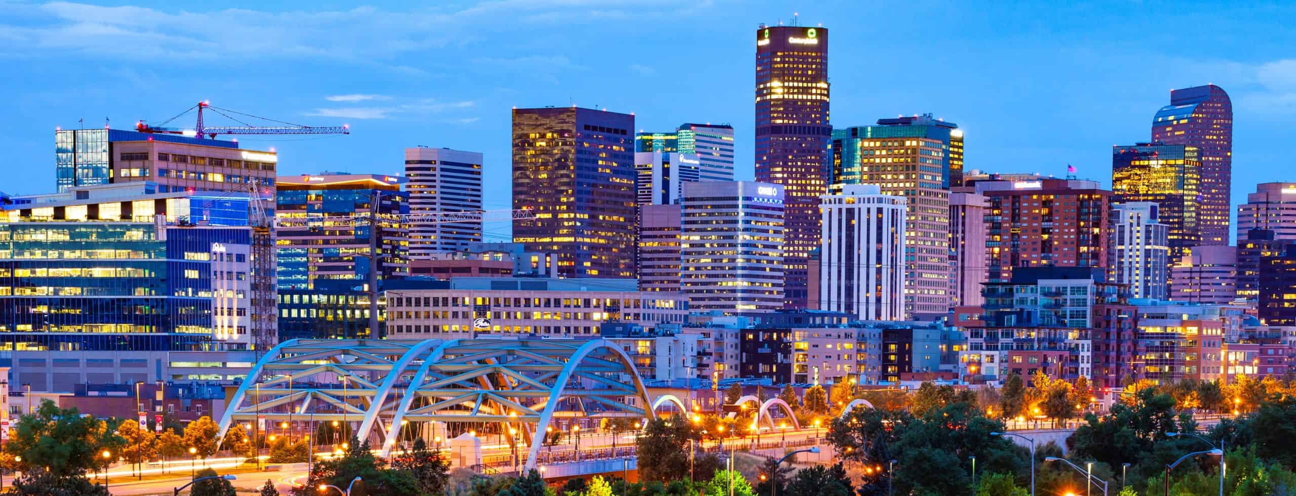 Modern city skyline at dusk with lit skyscrapers, a bridge in the foreground, a crane on the left, and lush greenery below.