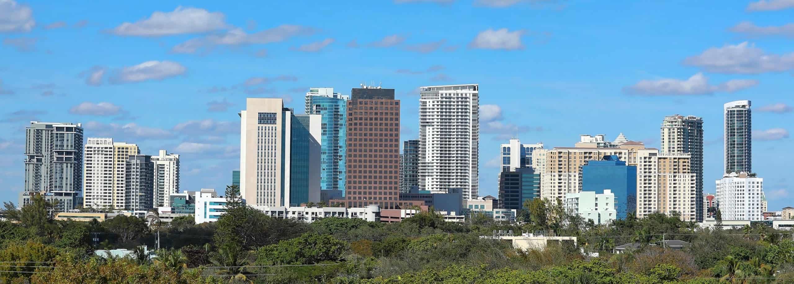 City skyline with modern skyscrapers under a blue sky, scattered clouds; green trees in the foreground create a striking contrast.