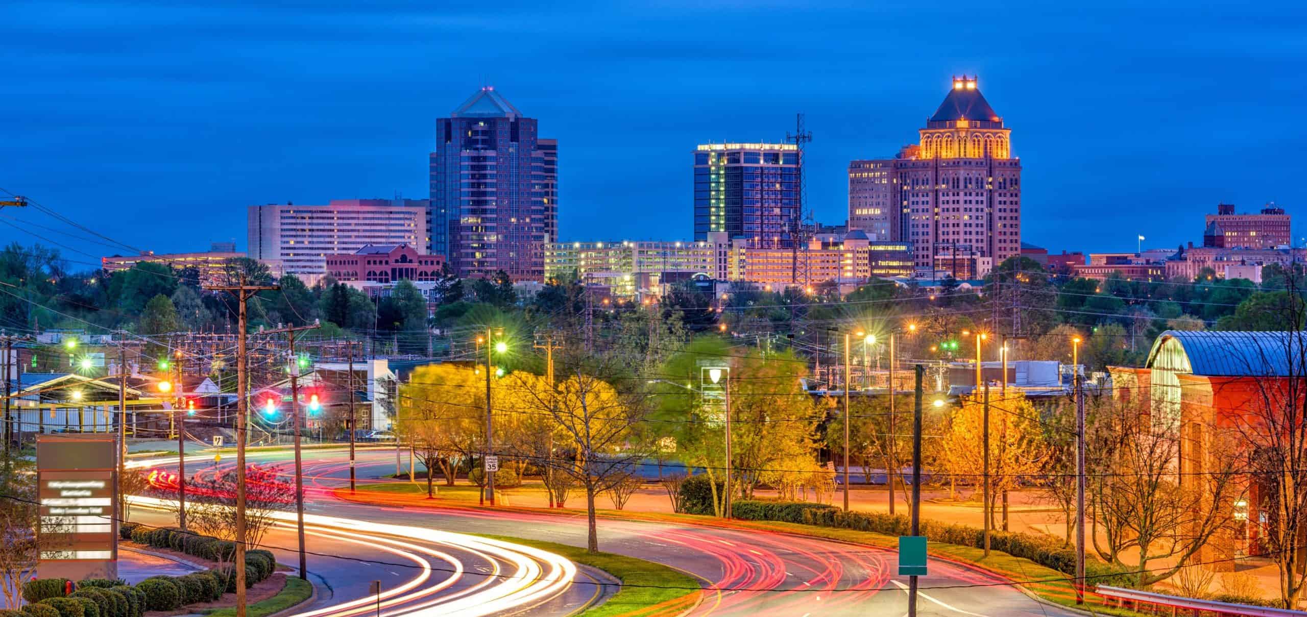 City skyline at dusk with lit buildings, a deep blue sky, car light trails on roads, trees, and streetlights adding ambiance.