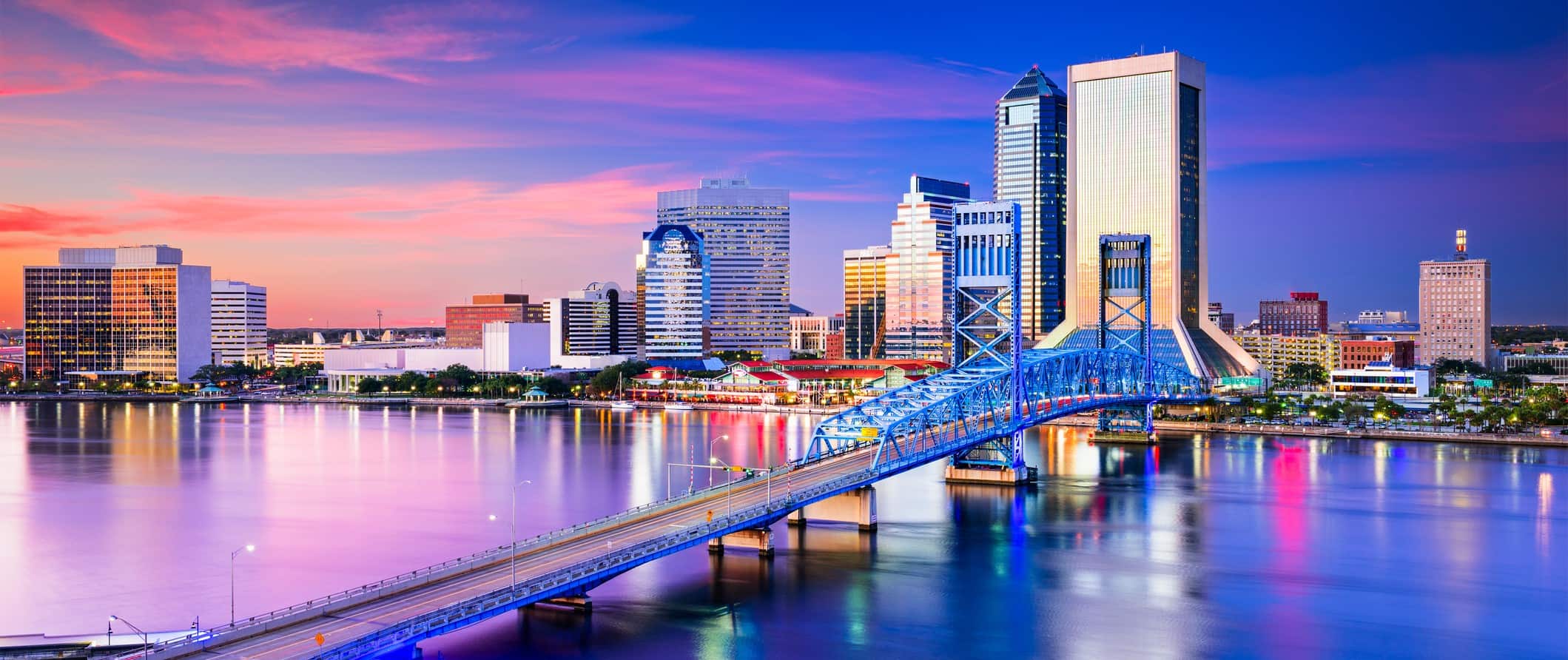 Colorful Jacksonville skyline at sunset: steel bridge, calm water, illuminated skyscrapers, vibrant pink and purple sky.