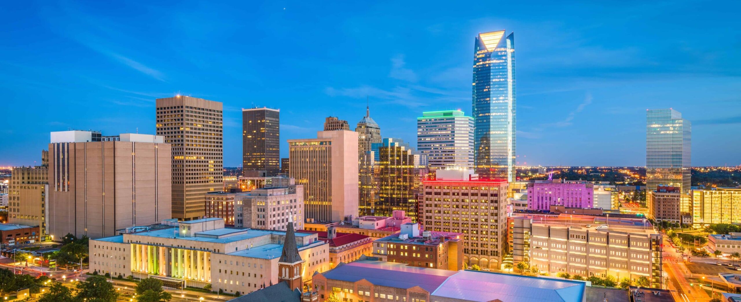 Vibrant city skyline at sunset: skyscrapers glow, tallest shines bright, colorful lights abound; clear sky shifts blue to deep.