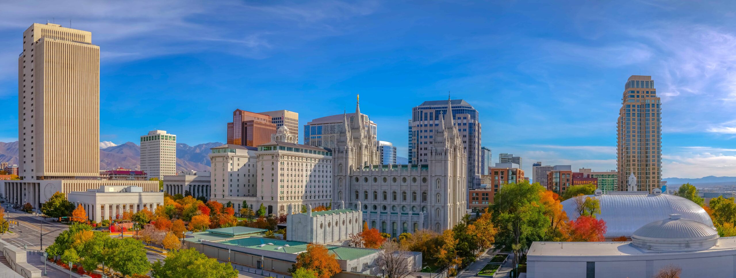 Cityscape with skyscrapers, historic architecture, and autumn trees; mountains in the background under a clear blue sky.