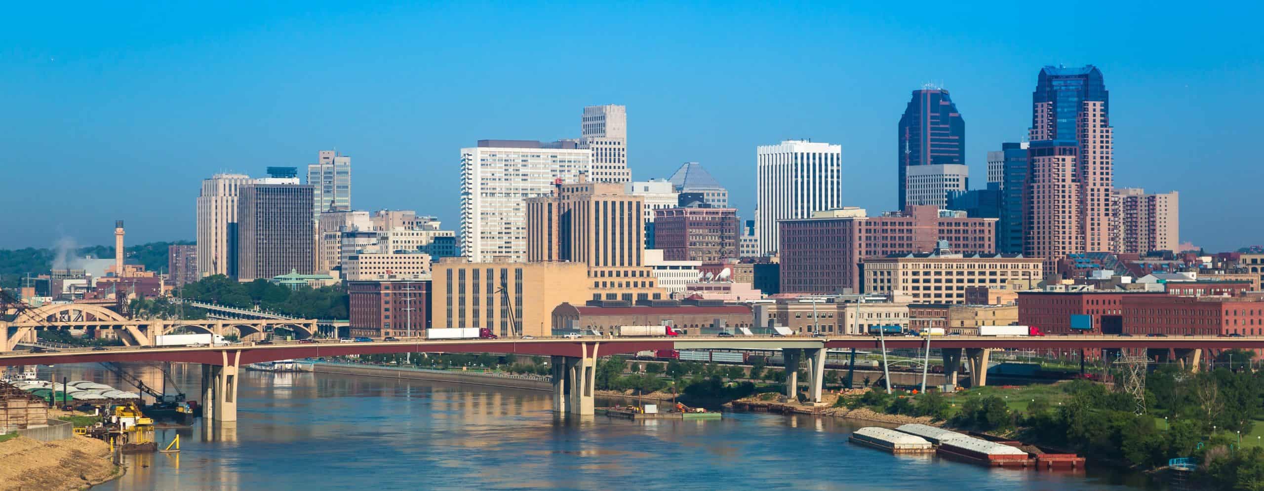 City skyline with tall buildings, clear blue sky. Arched bridge over river, lush greenery on banks, barges visible on water.