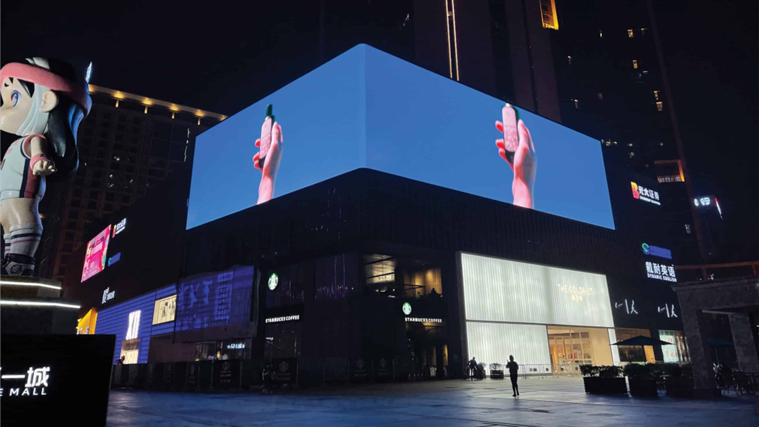 Billboard at night: hand with smartphone; few people, buildings, cartoon statue nearby.
