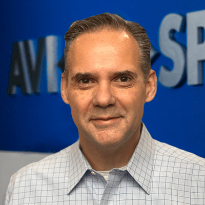 A man with short hair in a light checkered shirt stands before a blue background with partially visible text.