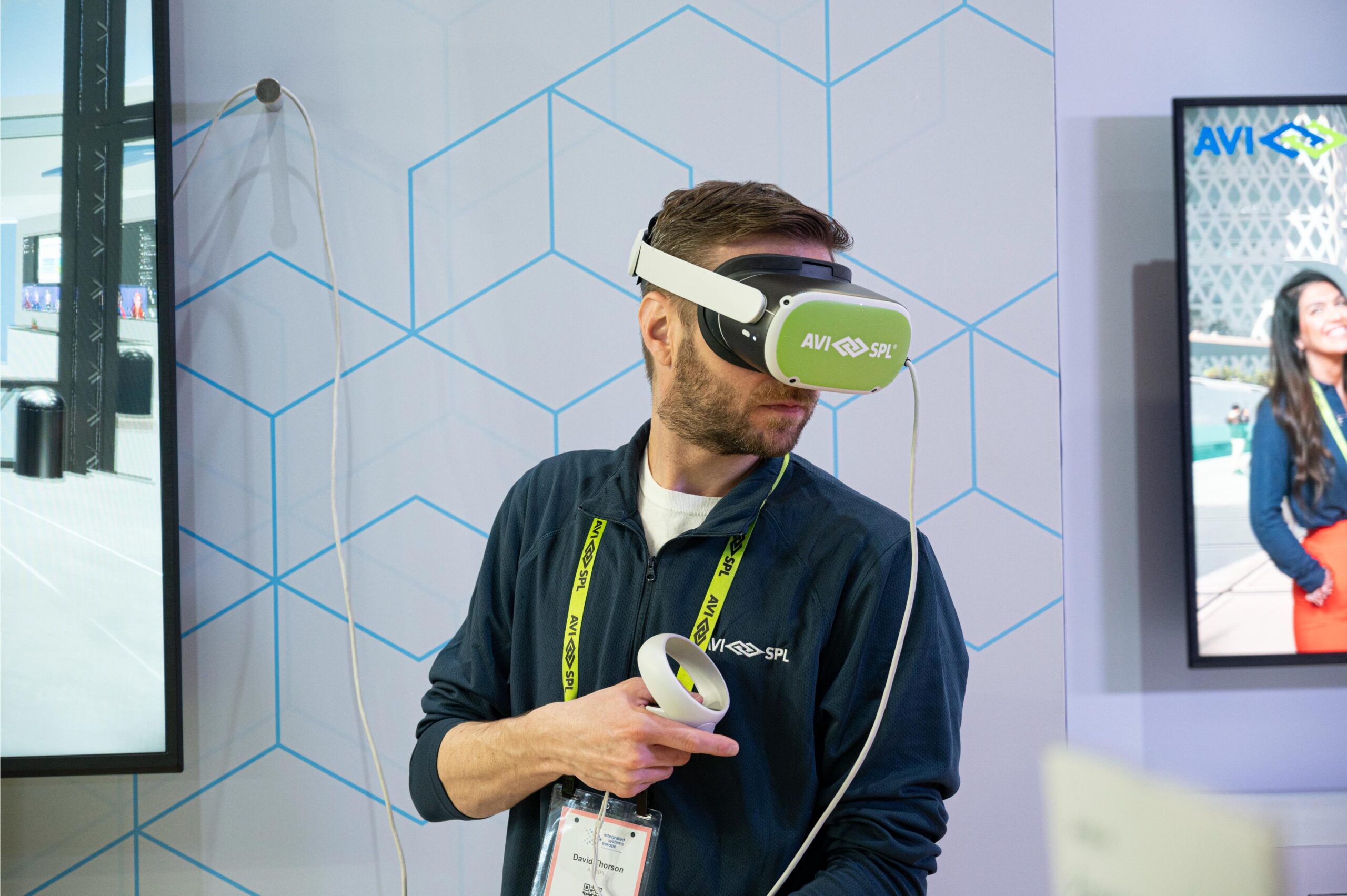 A person with a VR room design headset and controller, badge around neck, in front of screens displaying digital content; geometric background.