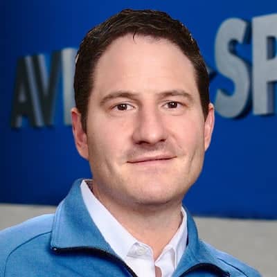 A short-haired person in a blue jacket and white shirt stands before a backdrop with partially visible blue and gray text.