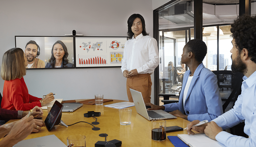 A diverse team meets with one presenting and screens showing charts. Laptops and papers fill the table, indicating business talks.