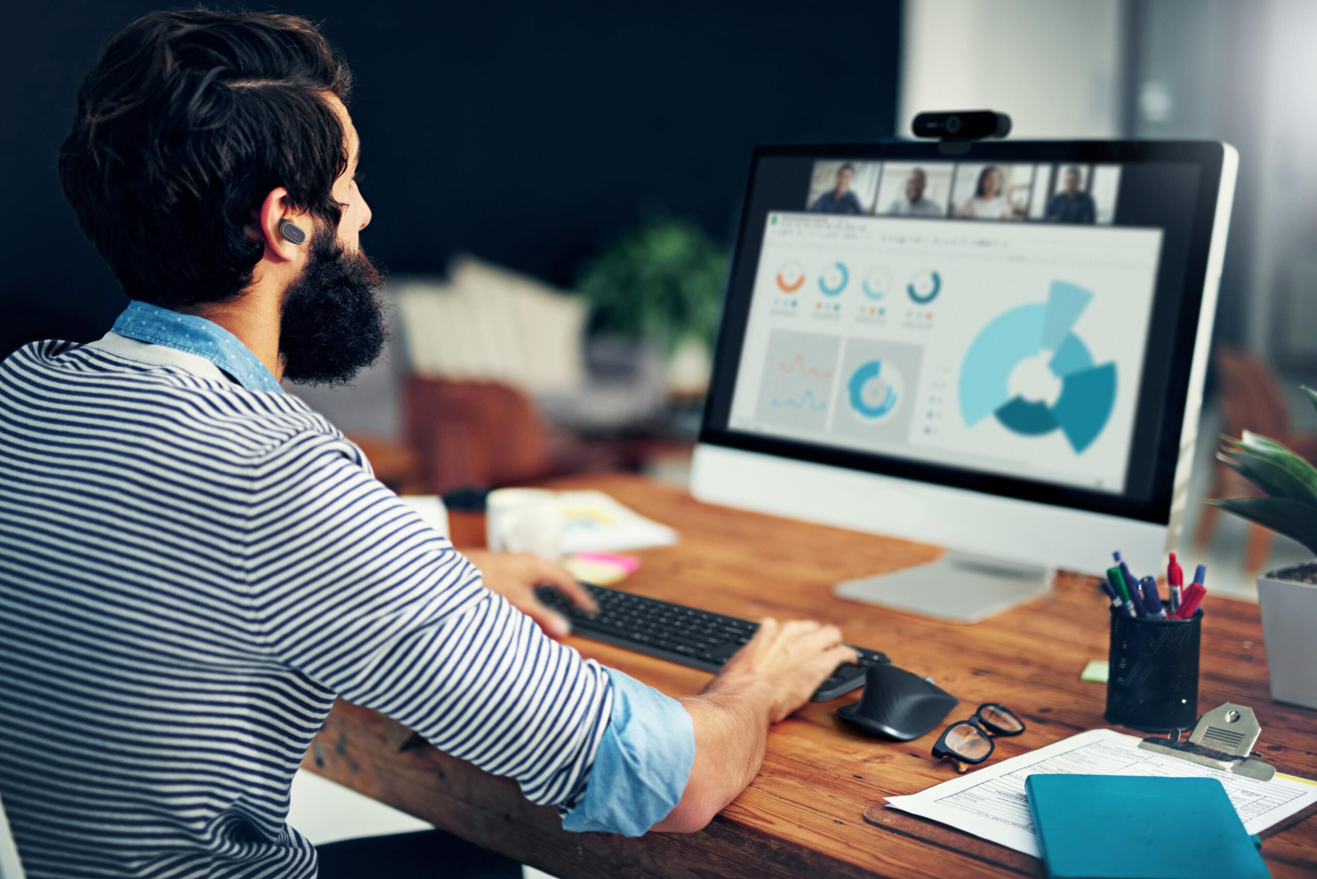 A bearded person at a desk views charts on a computer, wearing earbuds. A video call with 4 people is on screen; desk has plants and papers.