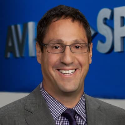 Person with glasses, gray suit, checkered shirt smiling; stands before blurred background with partial AVI and SP visible.