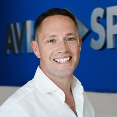 Smiling person in a white shirt stands before a blue wall with partially visible white text.