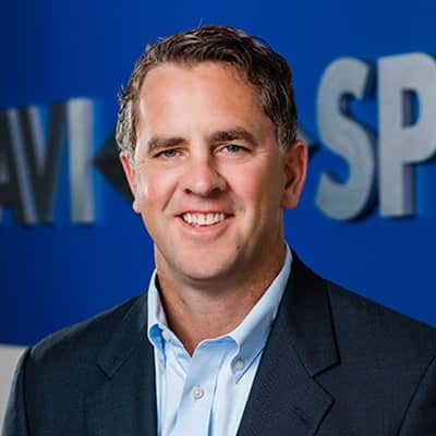 Smiling man in a suit with a blue shirt stands before a blurred sign with a blue background.