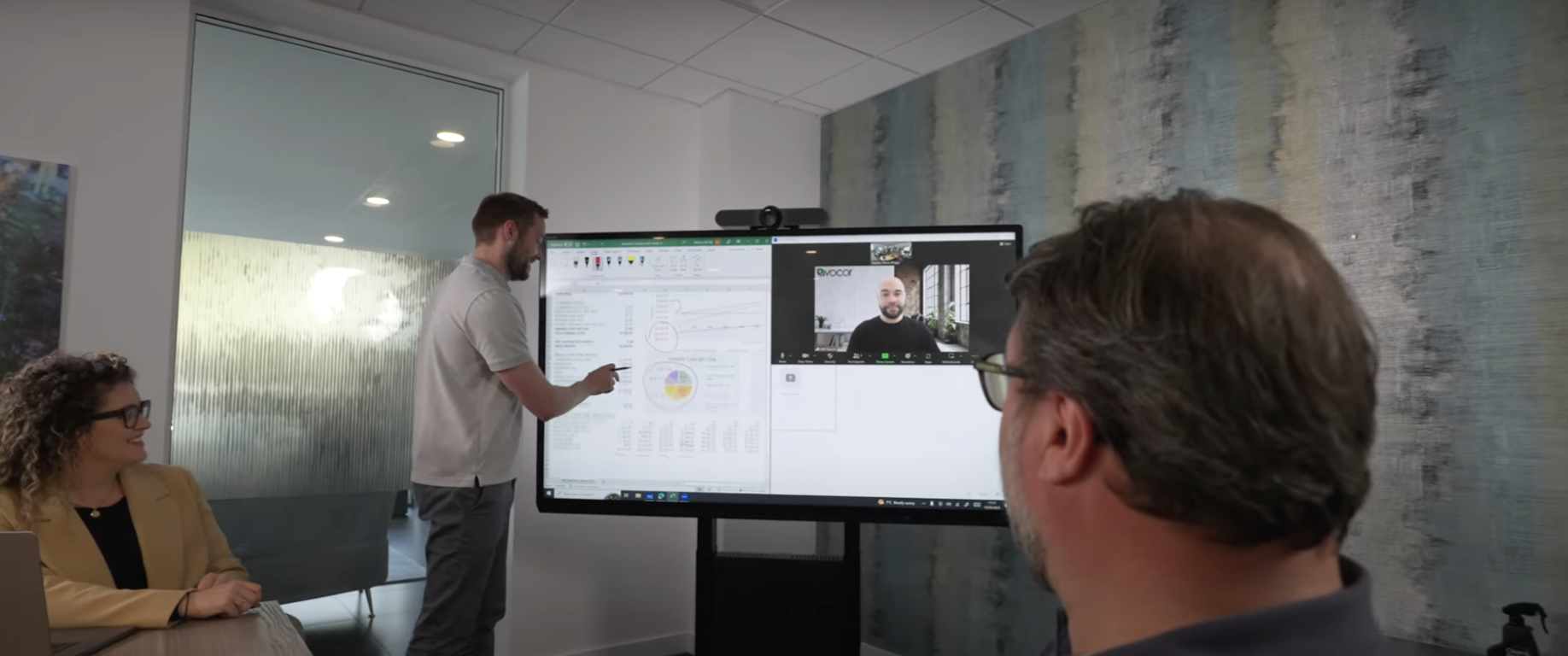 Man writes on interactive screen with charts; woman seated, man with glasses watch. Video call displayed.