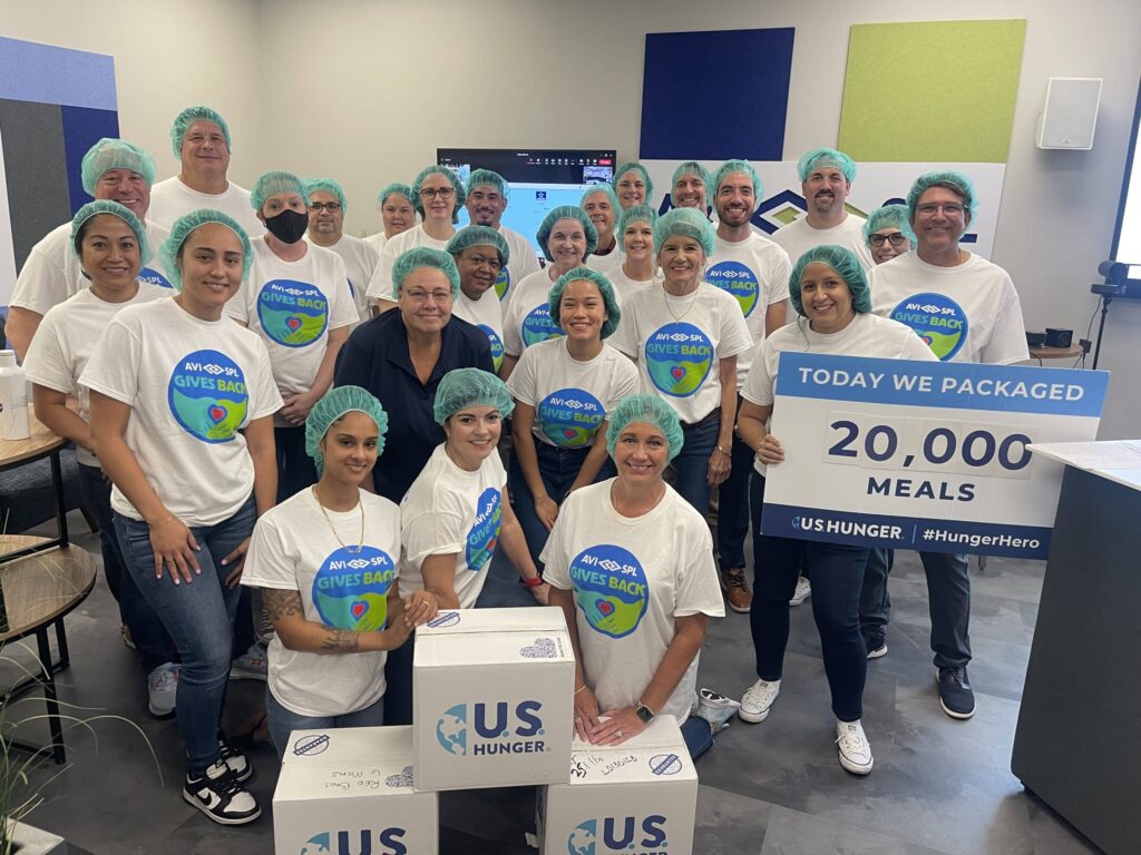 Group in hairnets and Gives Back shirts with US Hunger boxes, sign reads Packaged 20,000 meals today. Smiles highlight teamwork.