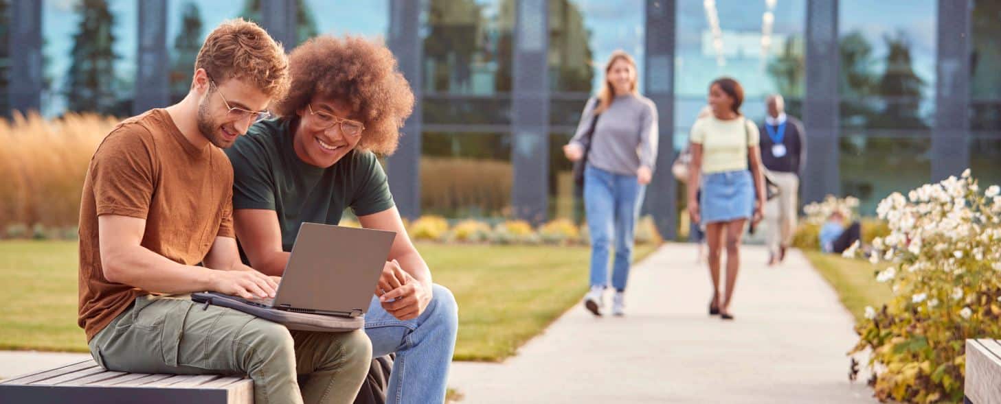 Two people sit smiling on a bench with a laptop; three walk by in an urban green space, modern glass building in the background.