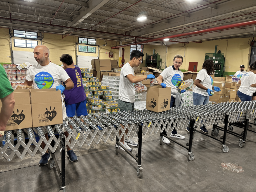 Volunteers in gloves pack food into boxes on a conveyor belt, wearing matching white shirts; stacks of goods sit behind them.