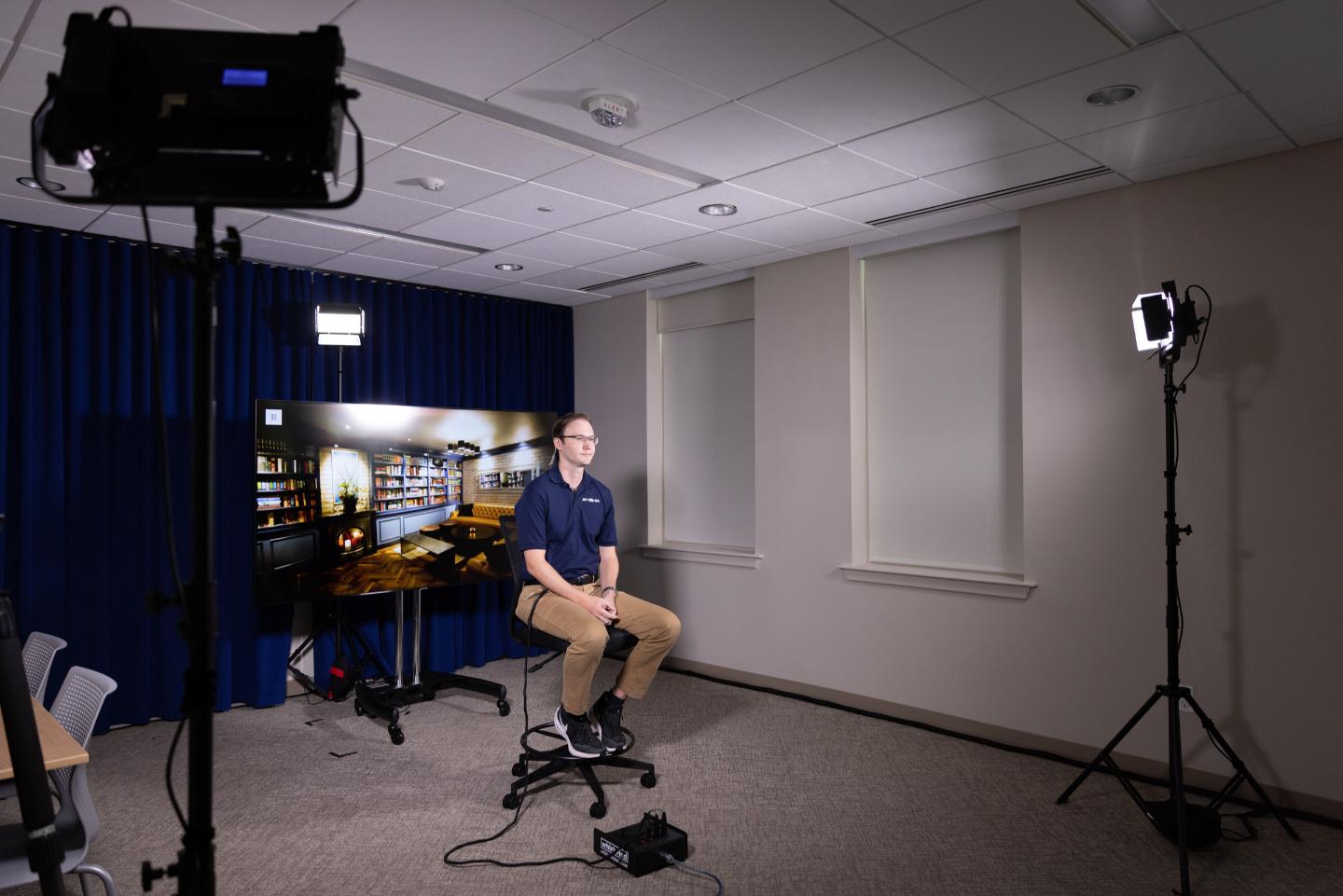 Man on stool in lit room, virtual bar backdrop, navy shirt & khakis, neutral expression. White walls & ceiling lights.