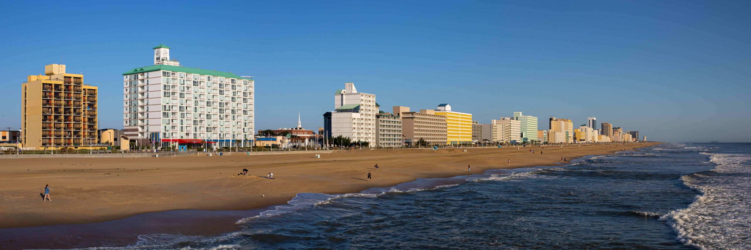Panoramic beach view: gentle waves, high-rise hotels, sunny sky, people strolling.