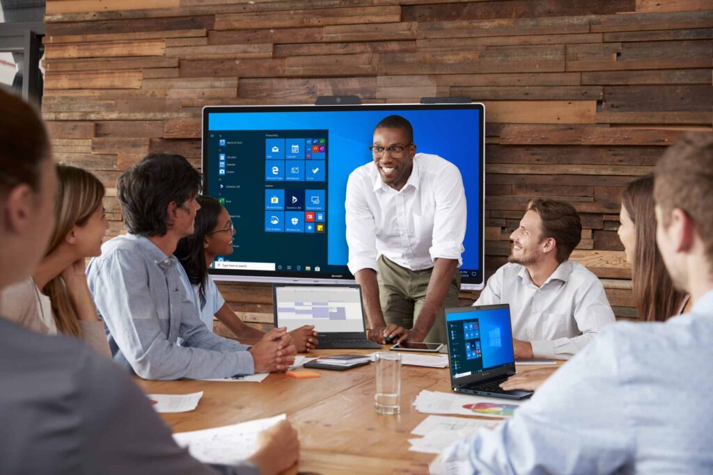 Man presents at meeting; colleagues seated with laptops, screen displays interface, rustic wooden wall background.