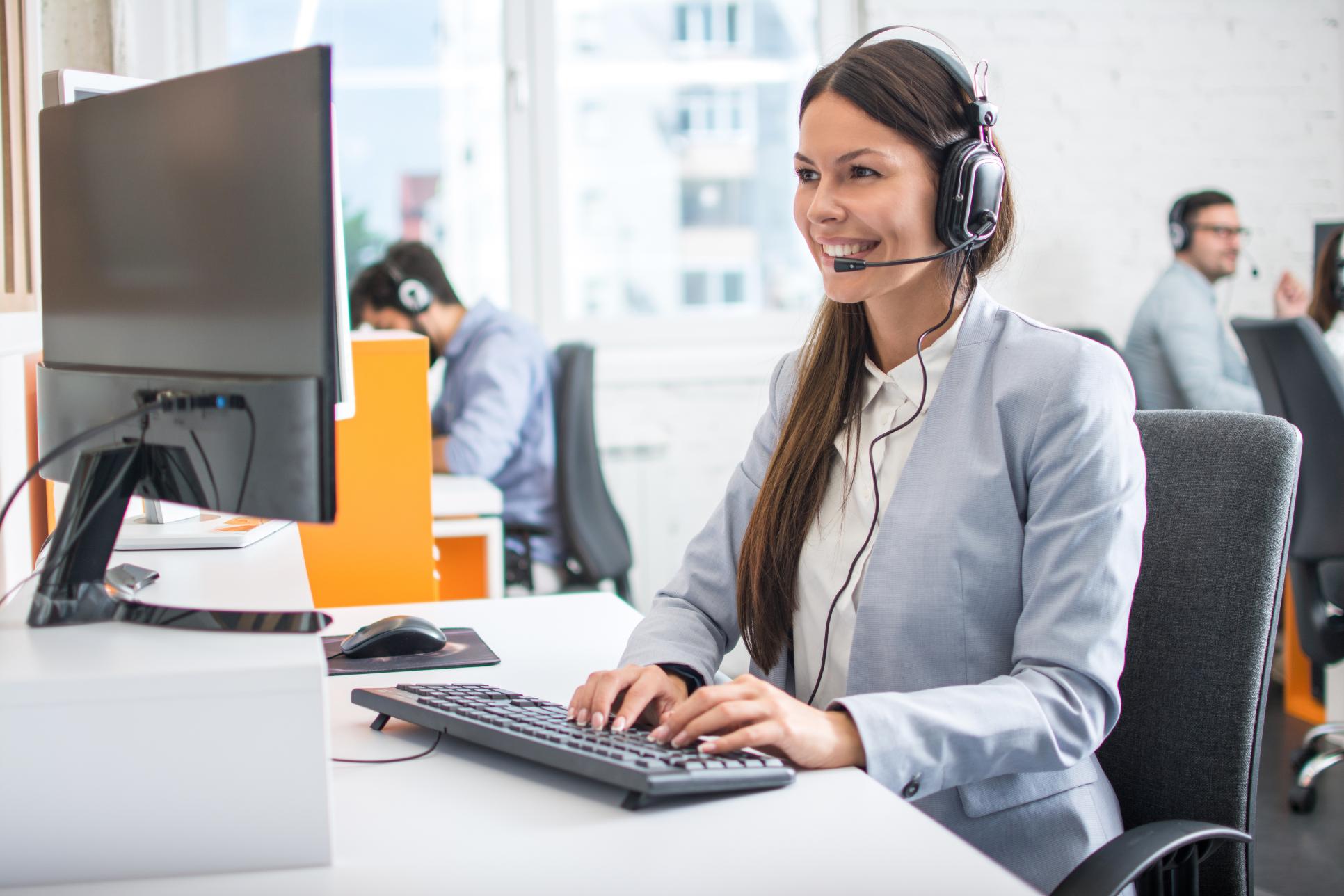 Woman in gray blazer and headset smiles typing at a computer; others in headsets focus on screens in the background.