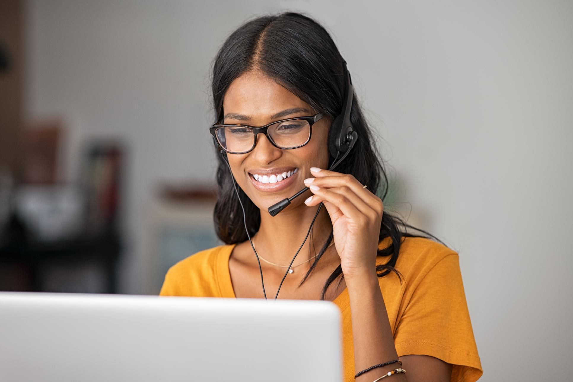 A woman with long hair, glasses, and a headset smiles at a laptop. She wears a yellow top; the background is blurred.