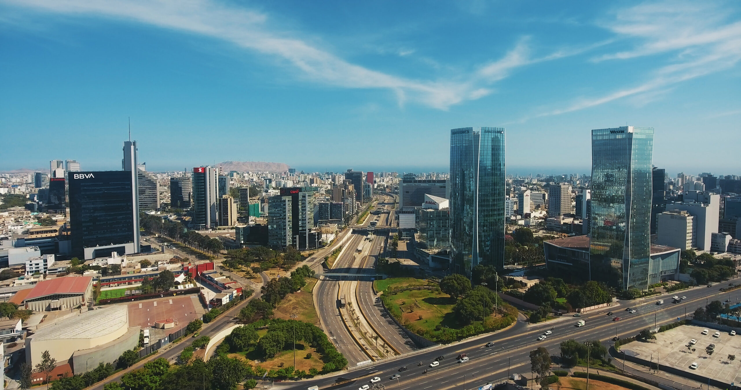 Cityscape aerial view: two glass skyscrapers, highway, diverse buildings, trees, mountains, blue sky with scattered clouds.