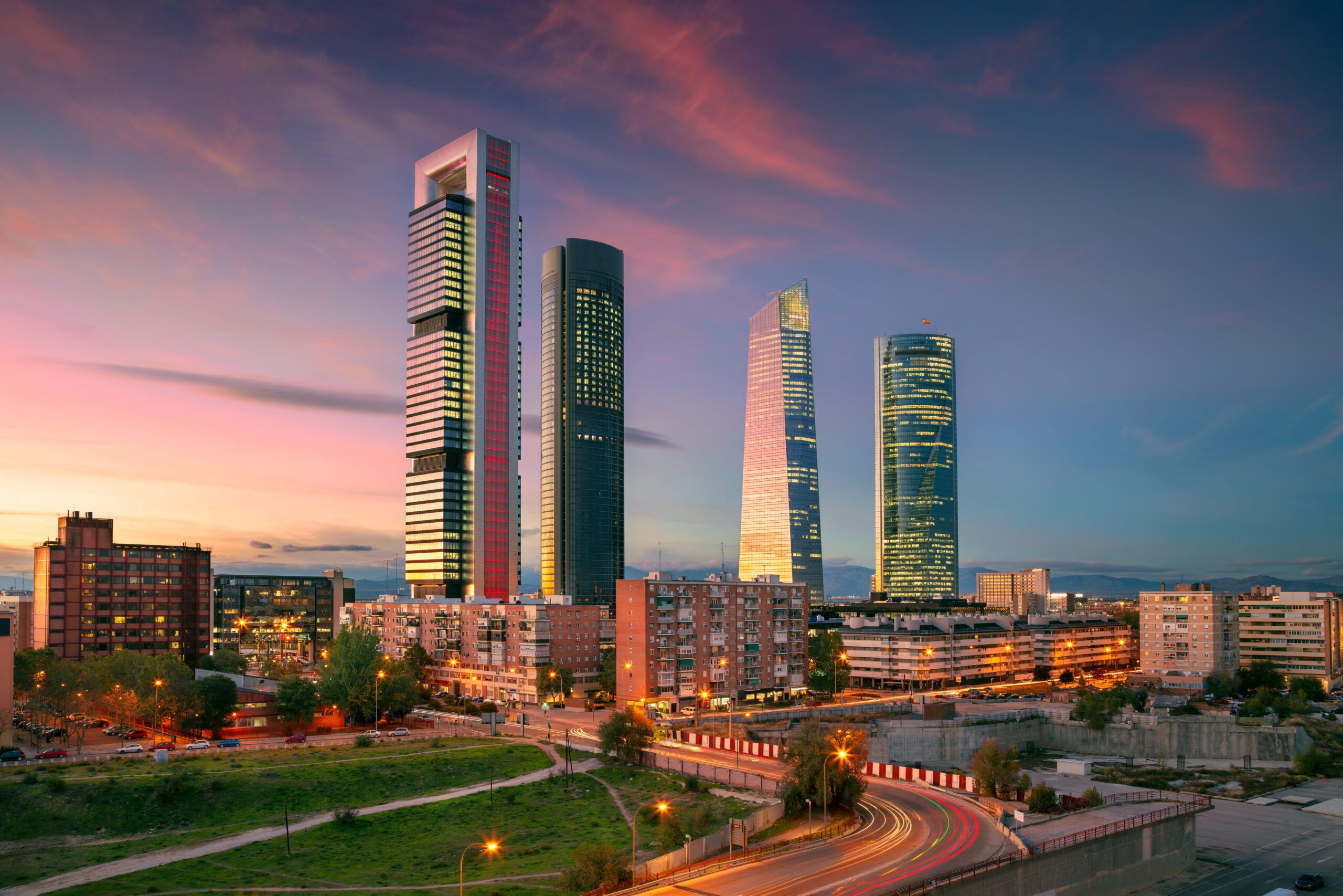 Cityscape at dusk: four lit skyscrapers, pink-orange sky; foreground of homes and roads with car lights.