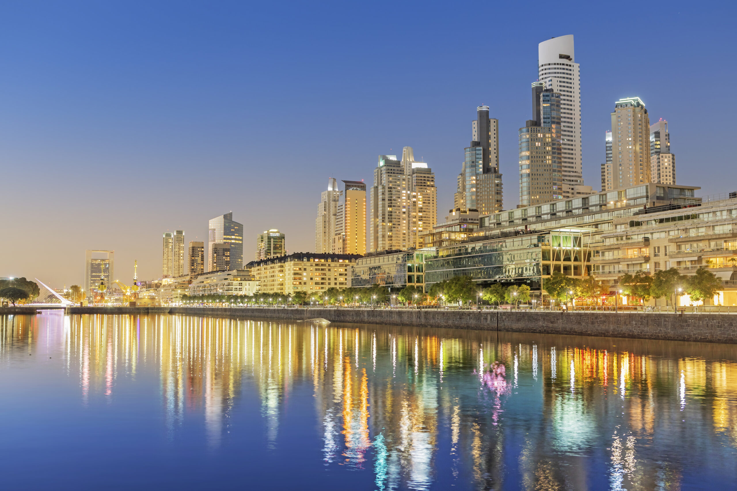 Buenos Aires skyline at dusk with skyscrapers reflecting in calm river, city lights aglow, and pedestrian bridge on the left.
