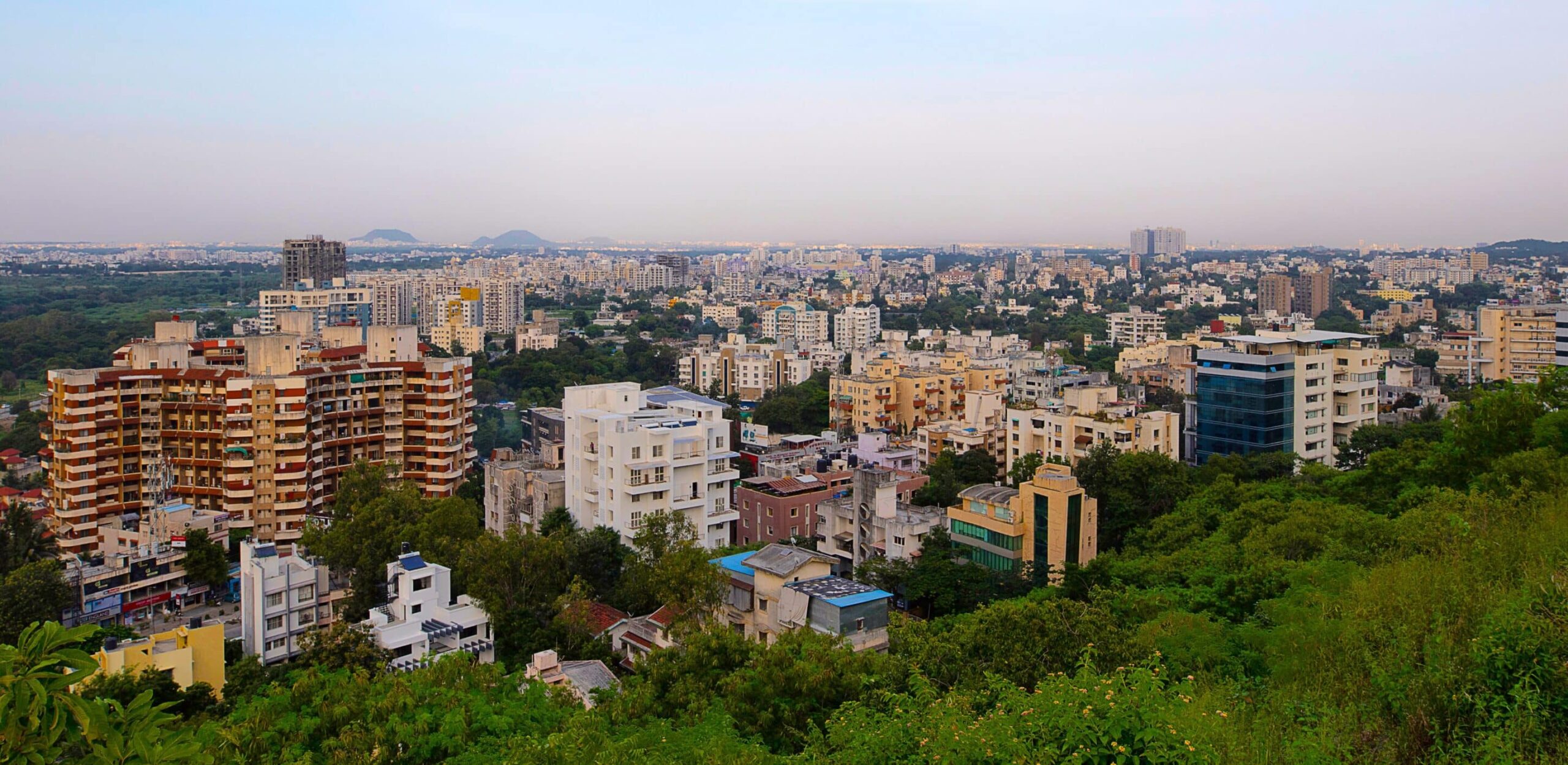 Cityscape with residences and offices amid greenery under clear skies; distant hills peek from the horizon.