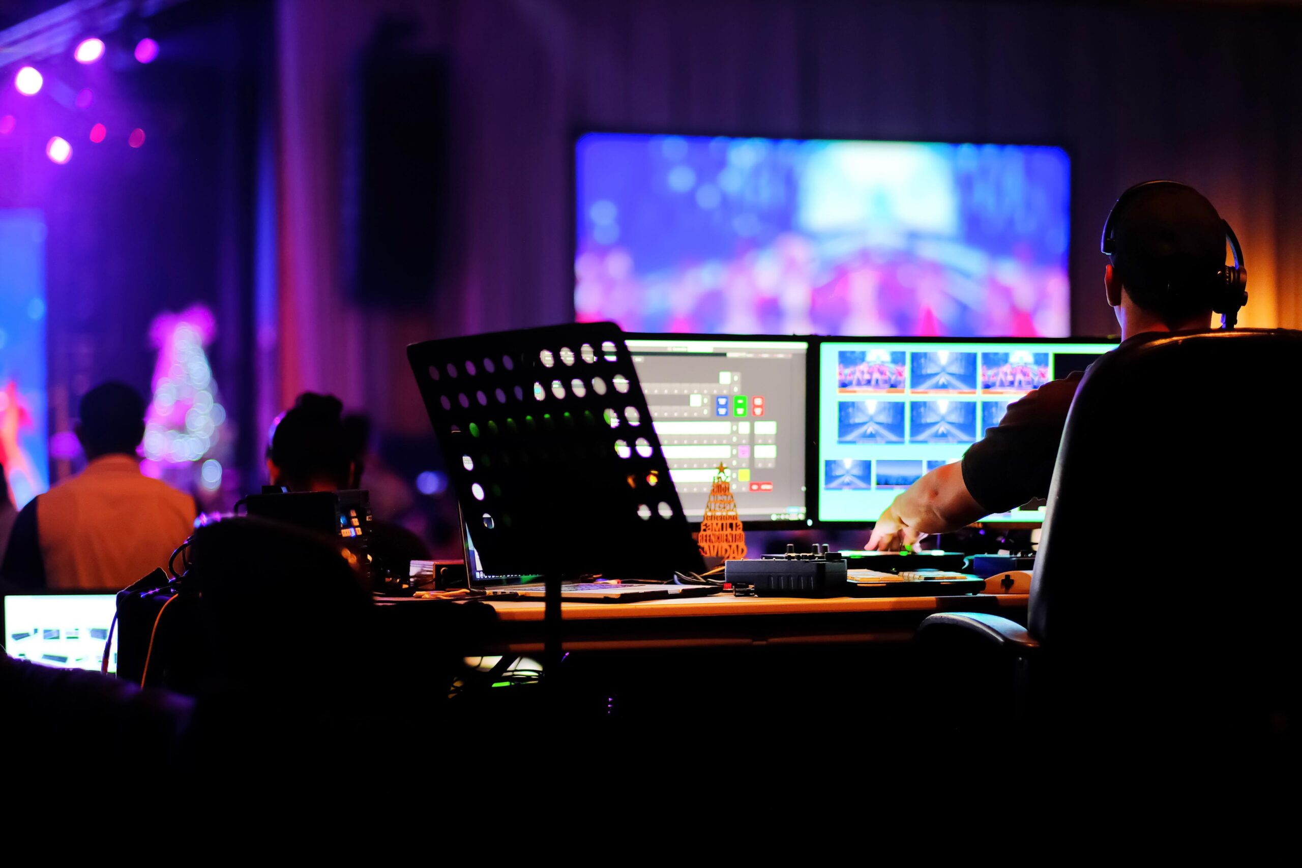 An AV mixer operates at an event with screens, purple lights, a performer on stage, and a Christmas tree decoration on the desk.
