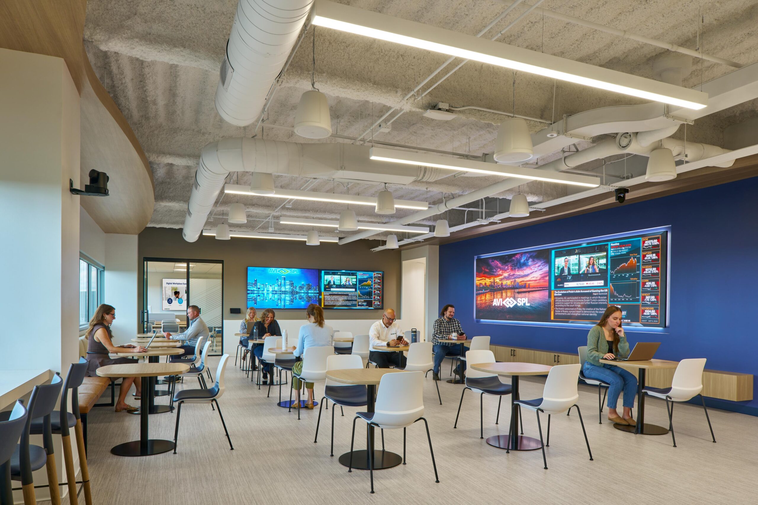 Modern office lounge: people working on laptops, discussing. Digital screens on walls, white furniture, exposed ducts.