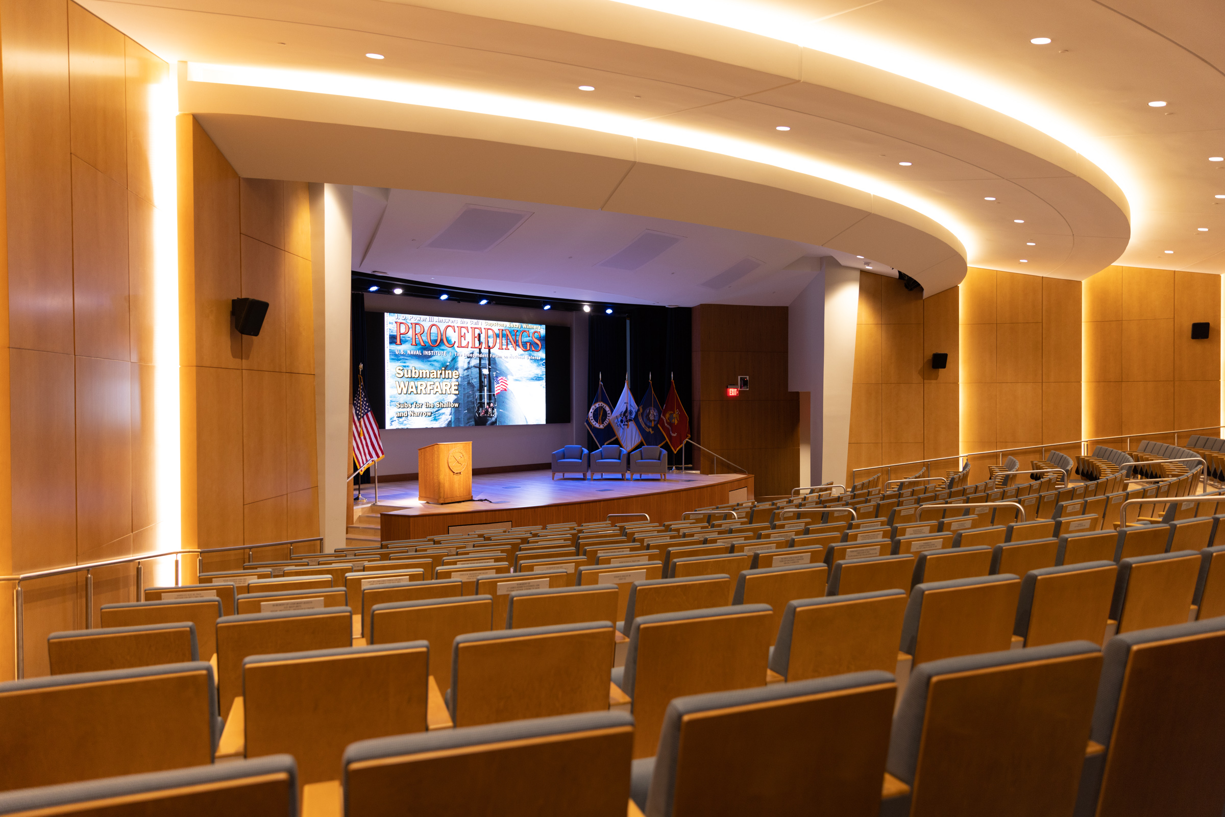 Empty auditorium with wooden seats facing a stage, Proceedings on screen, flags, soft lighting, wood-paneled walls and ceiling.